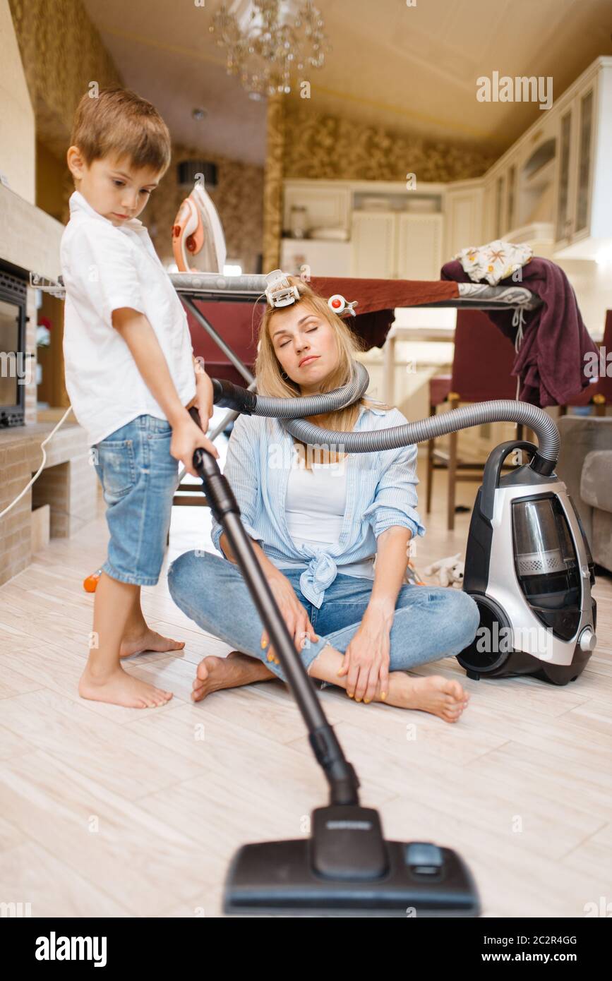 Un niñito envuelto el ama de casa con una manguera del aspirador. Mujer  realizando tareas domésticas en casa. Persona del sexo femenino con su hijo  juguetón engañando a arou Fotografía de stock -