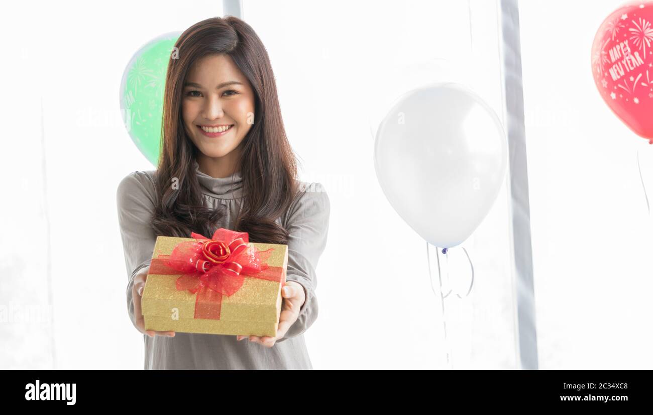 Una hermosa mujer asiática de 20-30 años sonríe alegremente y felizmente en una habitación blanca en su mano sosteniendo una caja de regalo dorada atada con un arco rojo como Foto de stock
