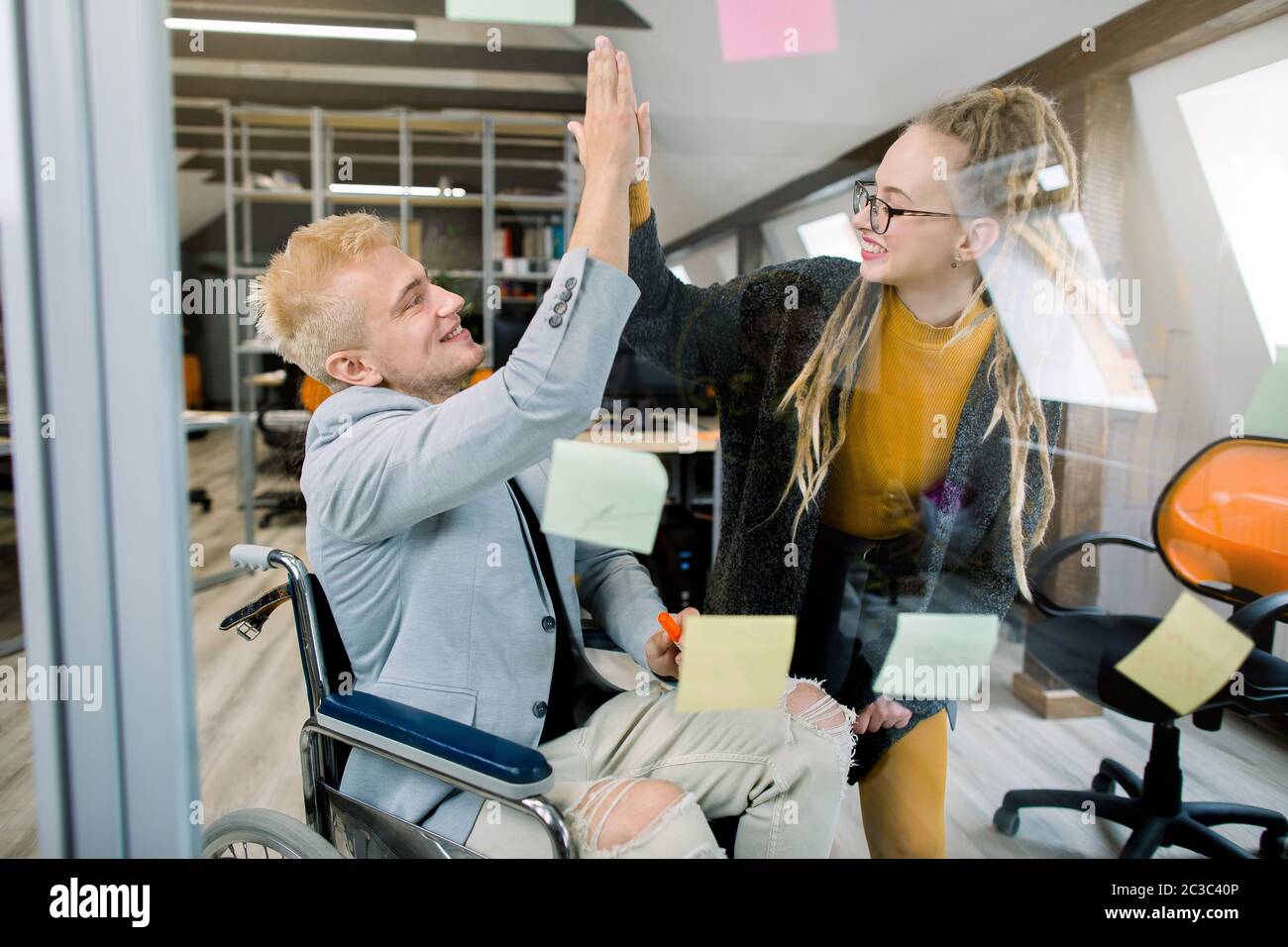 Concepto de negocio, personas con discapacidad en el trabajo. Feliz hombre joven exitoso en silla de ruedas dando un alto cinco a su cocolega bastante femenino con Foto de stock