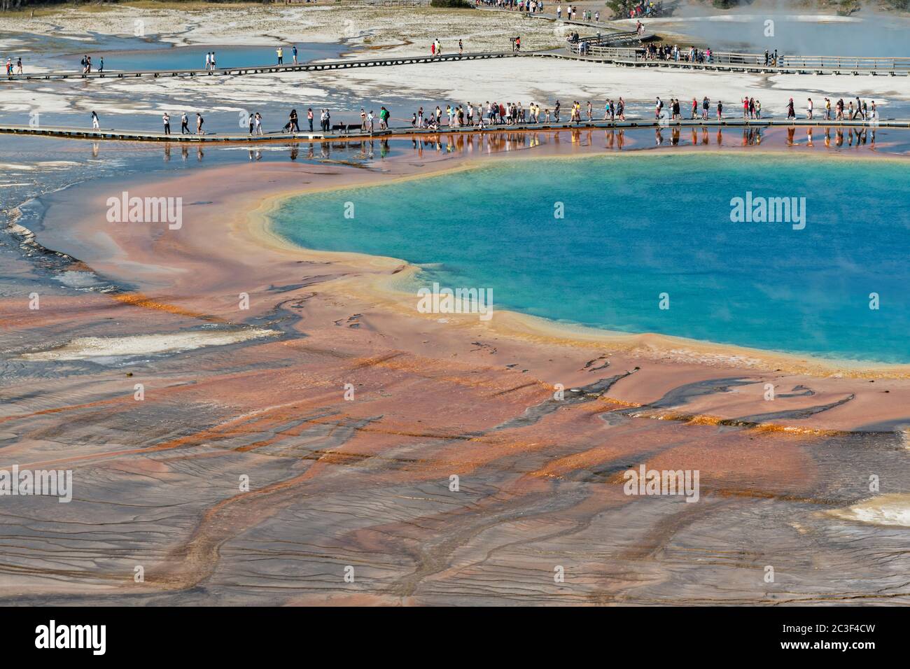 Los turistas caminan a lo largo del paseo marítimo en Grand Prismatic Spring, el manantial termal más grande del Parque Nacional Yellowstone y el tercero más grande del mundo. Grand Prismatic tiene aproximadamente 250 por 300 pies de tamaño, un promedio de 160 grados Fahrenheit y tiene hasta 160 pies de profundidad. Los colores brillantes alrededor de la primavera son de esteras de cianobacterias. El Grand Prismatic Spring es parte del Grupo Midway Geyser Basin Excelsior en Yellowstone, Wyoming. El Geyser Excelsior se puede ver detrás. Foto de stock