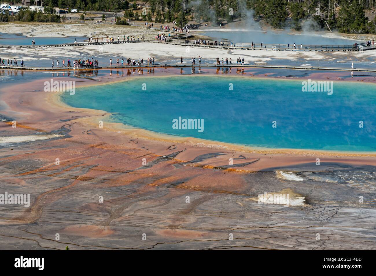 Los turistas caminan a lo largo del paseo marítimo en Grand Prismatic Spring, el manantial termal más grande del Parque Nacional Yellowstone y el tercero más grande del mundo. Grand Prismatic tiene aproximadamente 250 por 300 pies de tamaño, un promedio de 160 grados Fahrenheit y tiene hasta 160 pies de profundidad. Los colores brillantes alrededor de la primavera son de esteras de cianobacterias. El Grand Prismatic Spring es parte del Grupo Midway Geyser Basin Excelsior en Yellowstone, Wyoming. El Geyser Excelsior se puede ver detrás. Foto de stock