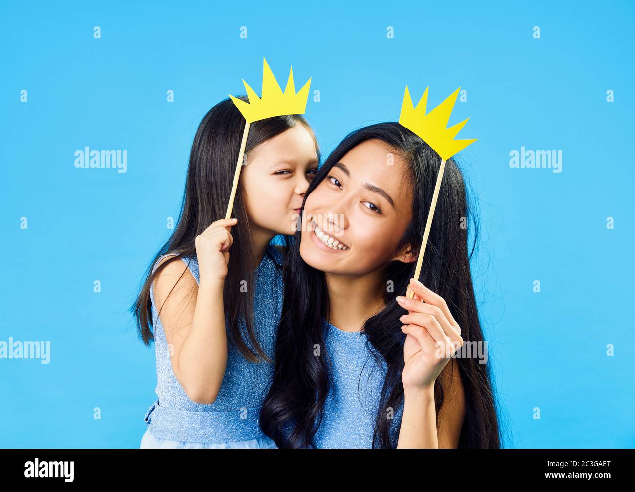 Linda hija besando a su feliz mamá joven sosteniendo la corona de papel en el palo sobre fondo azul divertirse juntos Foto de stock