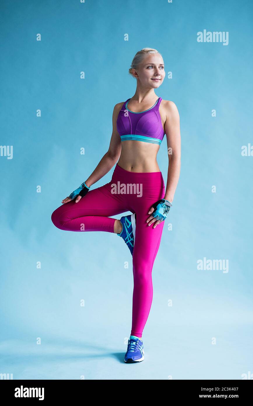 chica con ropa deportiva brillante se pone de pie en una pierna, fondo  azul, hermoso cuerpo deportivo curtido Fotografía de stock - Alamy