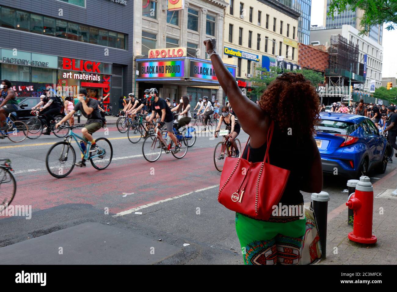 Nueva York, NY, 20 de junio de 2020. Una mujer con un puño levantado se encuentra frente al Teatro Apollo con 'Negro y orgulloso' en la marquesina mientras los manifestantes en ciclistas pasan. La protesta de la bicicleta fue un viaje de solidaridad de la materia de vidas negras que pidió justicia en una serie reciente de asesinatos de la policía estadounidense: George Floyd, Breonna Taylor, y muchos otros. El paseo en bicicleta fue organizado por el colectivo llamado Street Riders NYC. Varios miles de personas participaron en la manifestación itinerante viajando desde Times Square, Harlem y Battery Park. 20 de junio de 2020 Foto de stock
