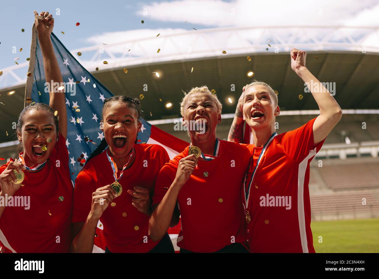 Equipo de fútbol americano femenino celebrando el campeonato. Las mujeres del equipo de fútbol gritando en el campo sosteniendo bandera nacional con confeti caen alrededor. Foto de stock