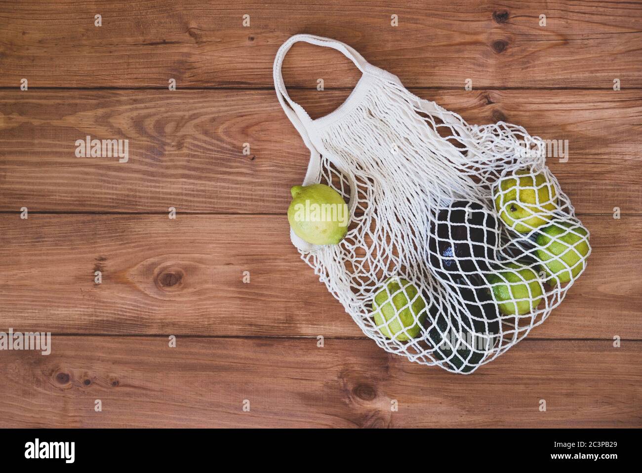 Bolsa de malla de algodón reutilizable ecológica con limones y aguacate sobre fondo rústico de madera. Foto de stock