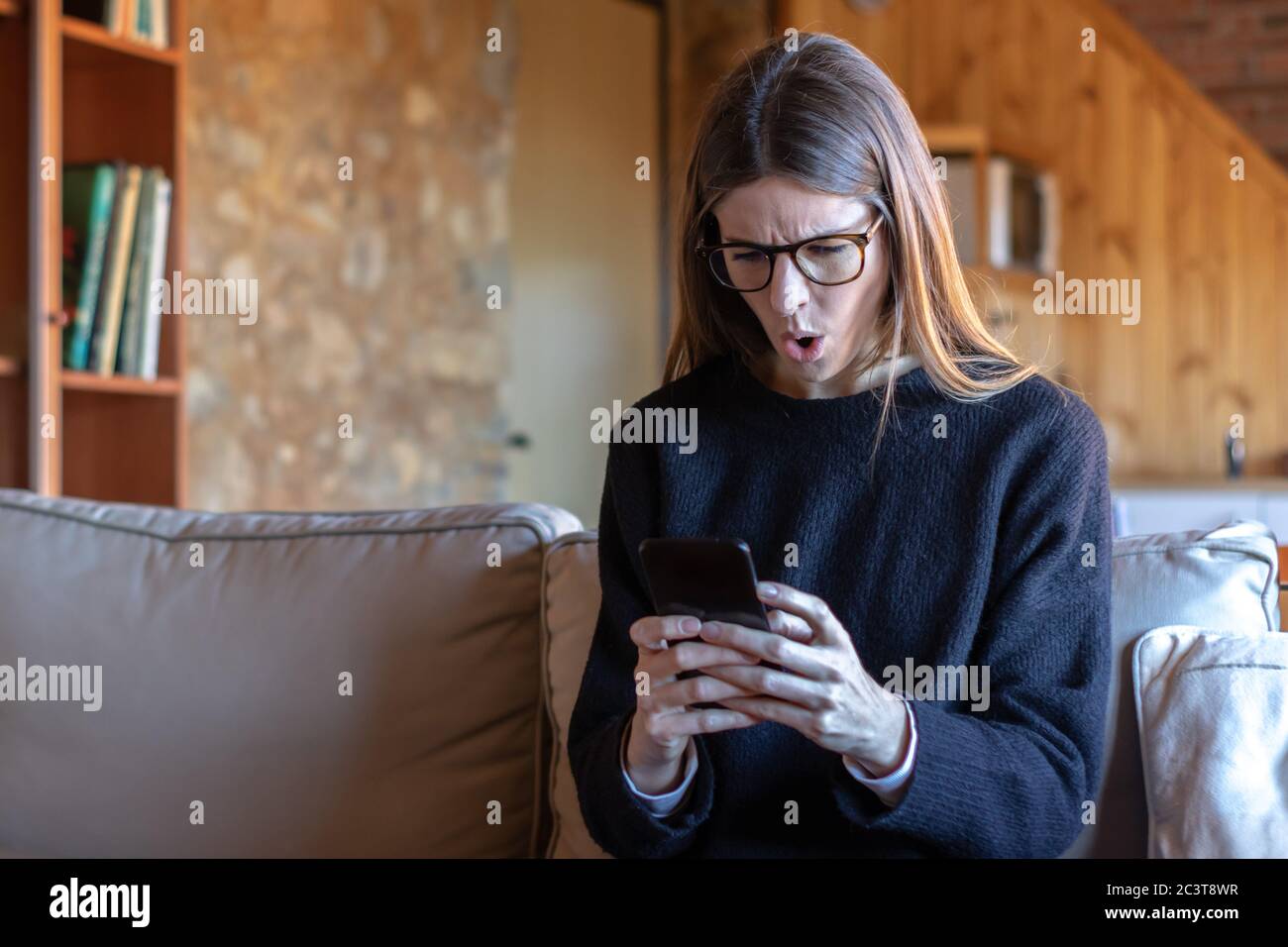 Sorprendida mujer joven morena con gafas escribiendo en el smartphone sentado en el sofá de casa usando un suéter negro Foto de stock