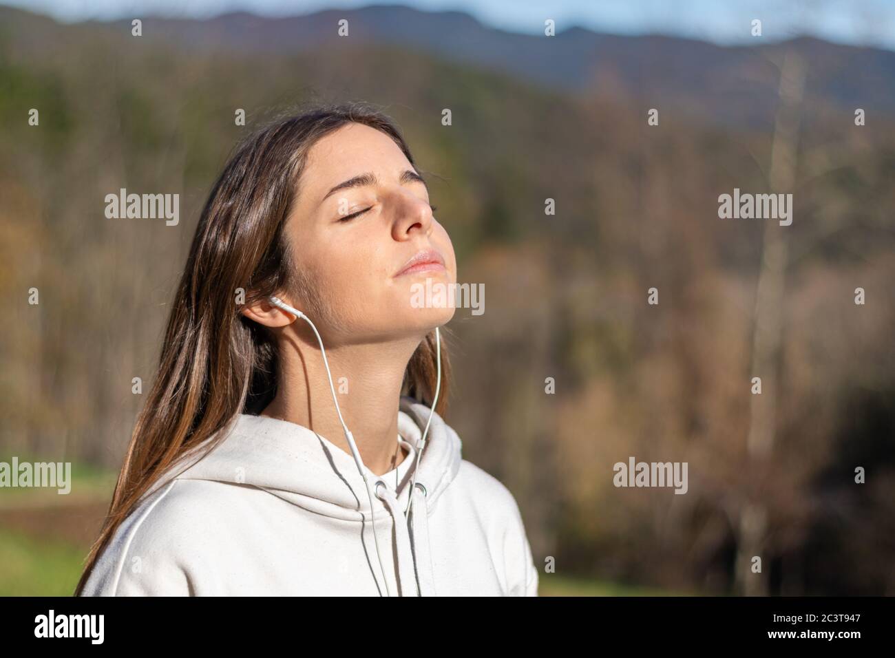 Una mujer joven y bonita respira aire fresco en el parque escuchando música  en los auriculares. Ella está usando una sudadera ligera y tiene un largo  cabello marrón Fotografía de stock -