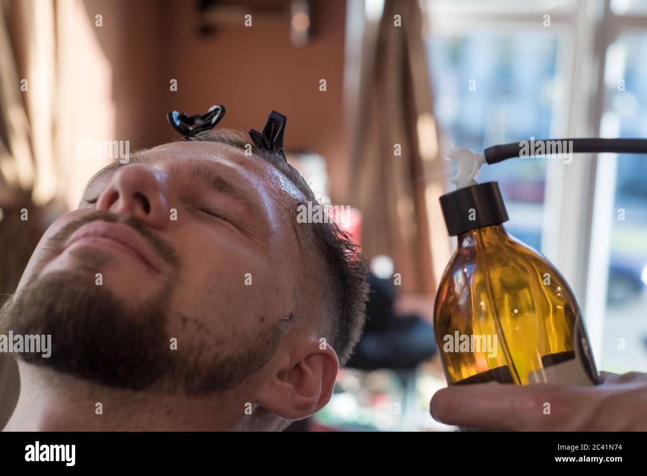 una peluquería rociando un bálsamo después de afeitarse la barba a un cliente de un joven barbudo de veinticinco años. A la peluquería Foto de stock