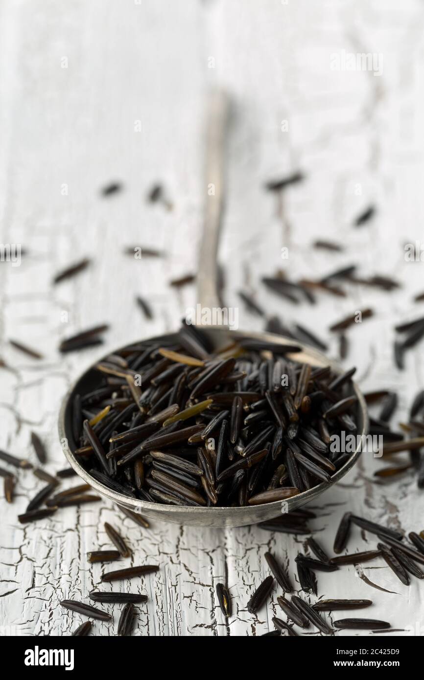 Montón de granos de arroz salvaje crudos, crudos y negros en cuchara de metal sobre fondo blanco de mesa rústica, enfoque selectivo Foto de stock