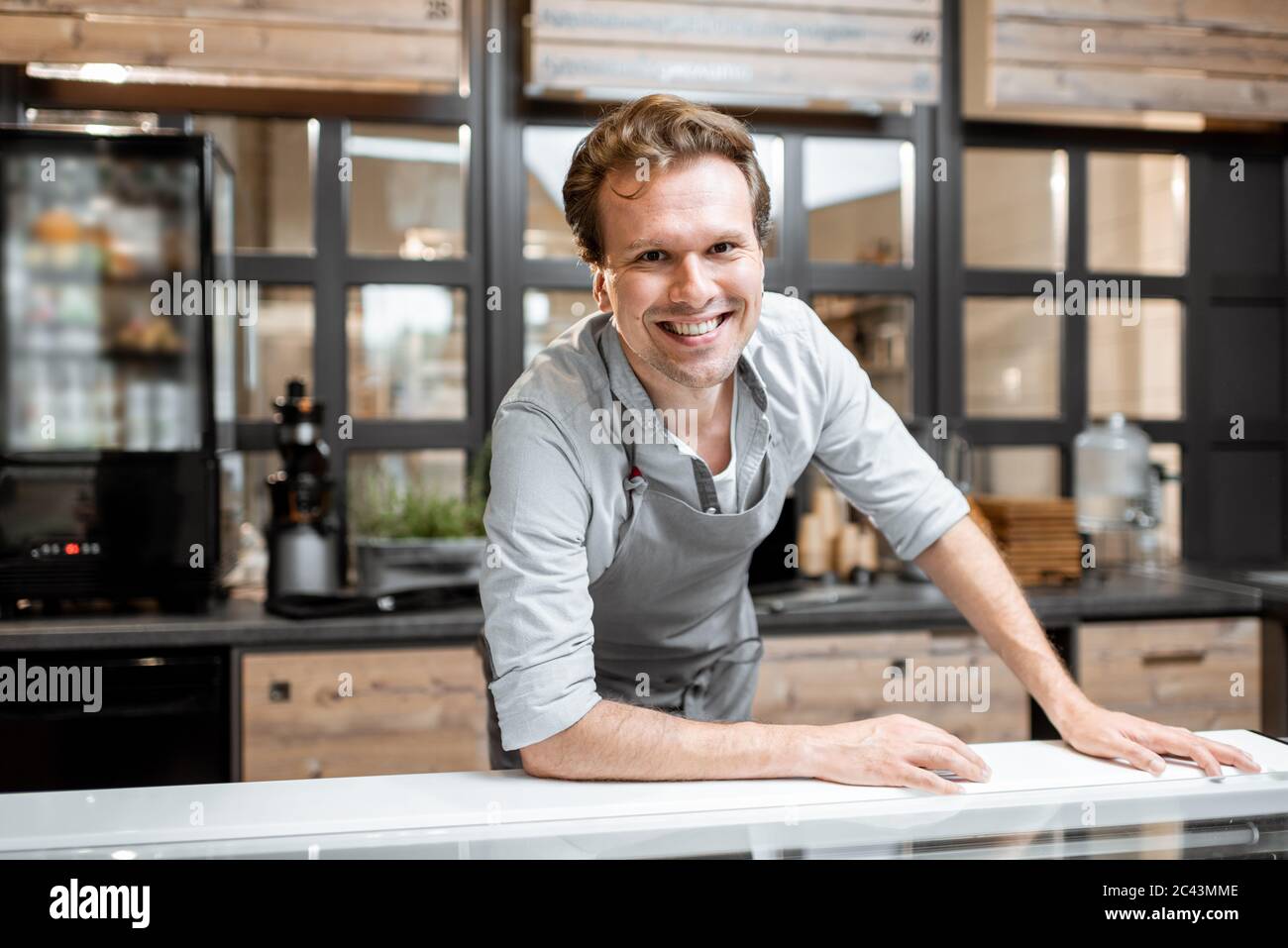 Retrato de un alegre vendedor en un delantal de pie detrás del mostrador de una pequeña tienda o cafetería. Concepto de pequeña empresa y trabajo en el campo de los servicios Foto de stock
