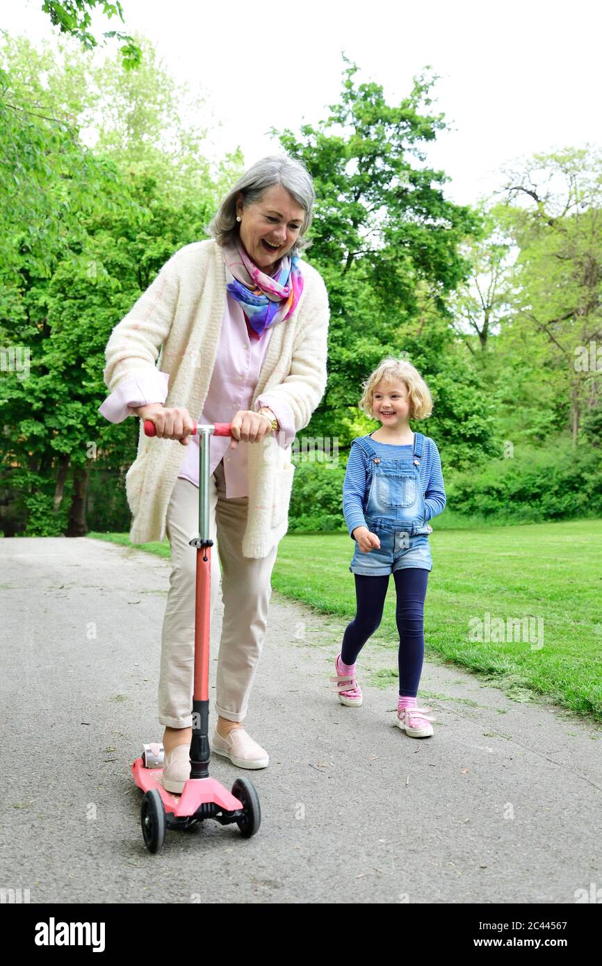 Alegre mujer mayor montar en scooter de empuje mientras nieta caminando en el parque Foto de stock