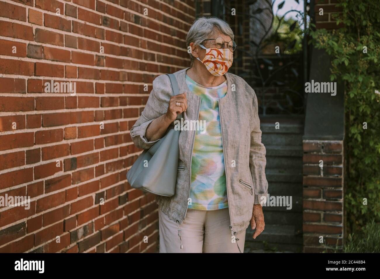 Mujer mayor jubilada usando máscara facial mientras caminaba por la pared de ladrillo durante la epidemia de coronavirus Foto de stock