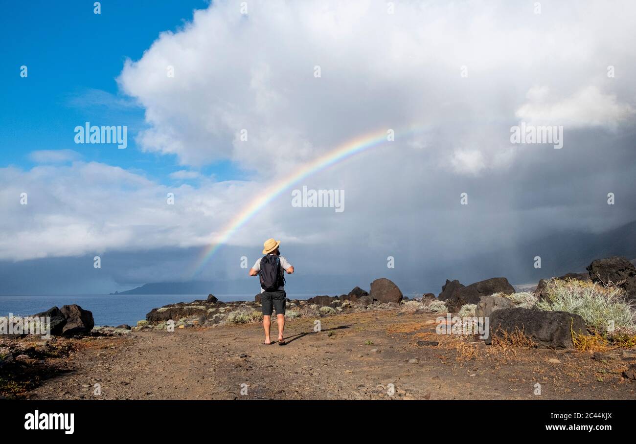 España, el Hierro, Hiker masculino admirando el doble arco iris sobre la zona costera rocosa Foto de stock