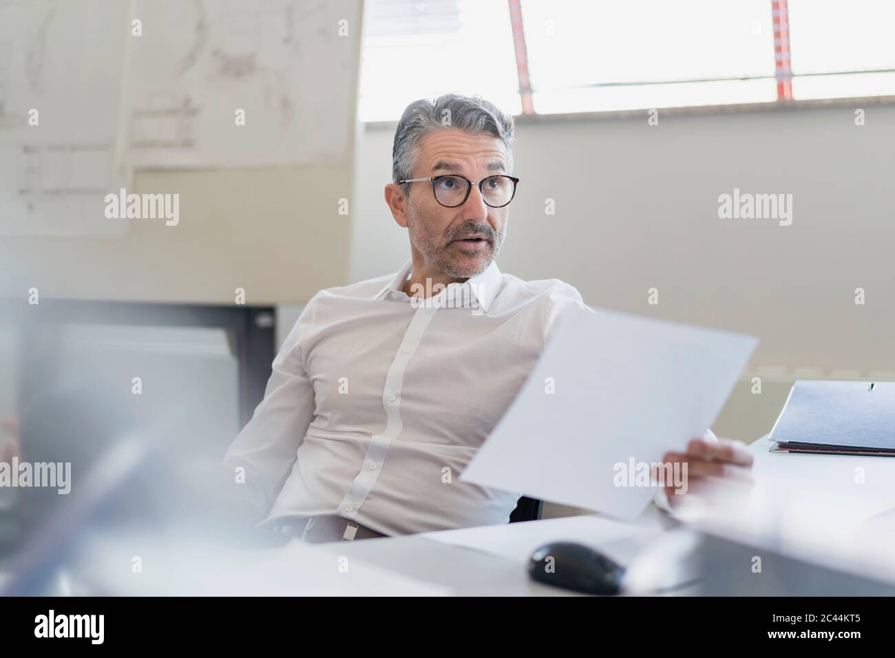 Hombre de negocios seguro mirando con cejas elevadas mientras se sienta con documentos en el escritorio de la oficina Foto de stock