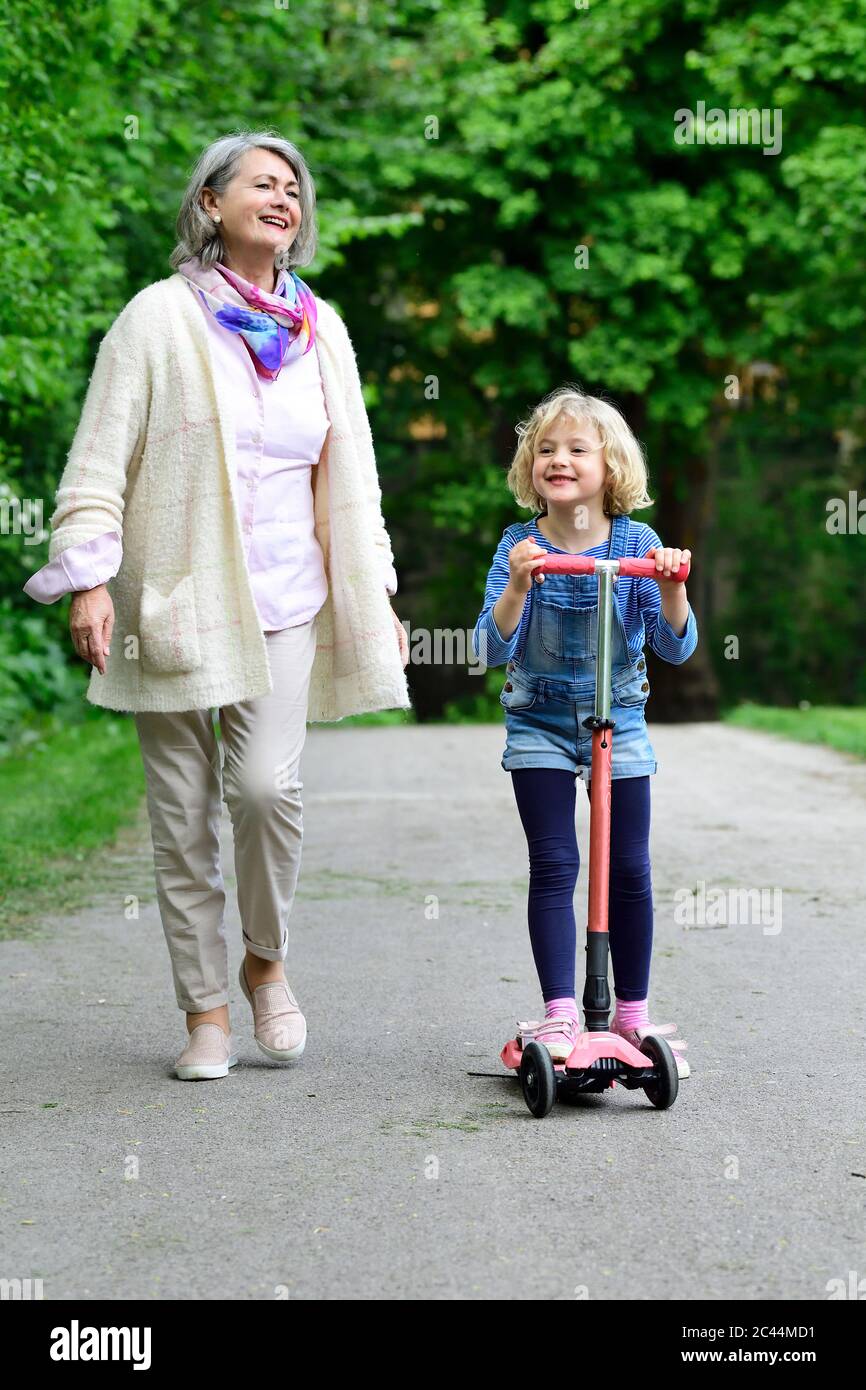 Feliz mujer mayor caminando con nieta montando scooter en el parque Foto de stock