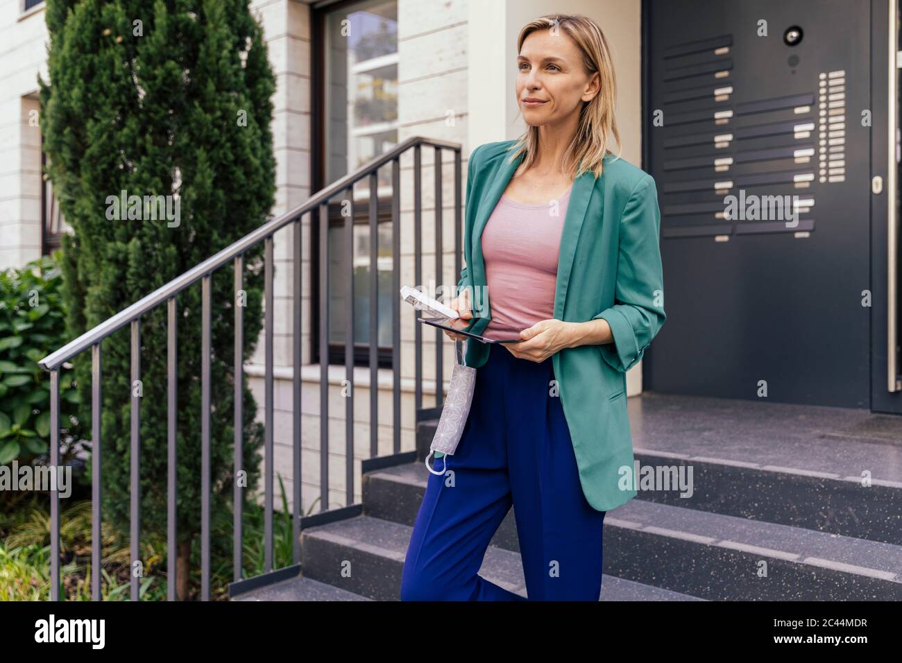 Mujer con tableta digital y máscara en la mano caminando por las escaleras de la casa Foto de stock