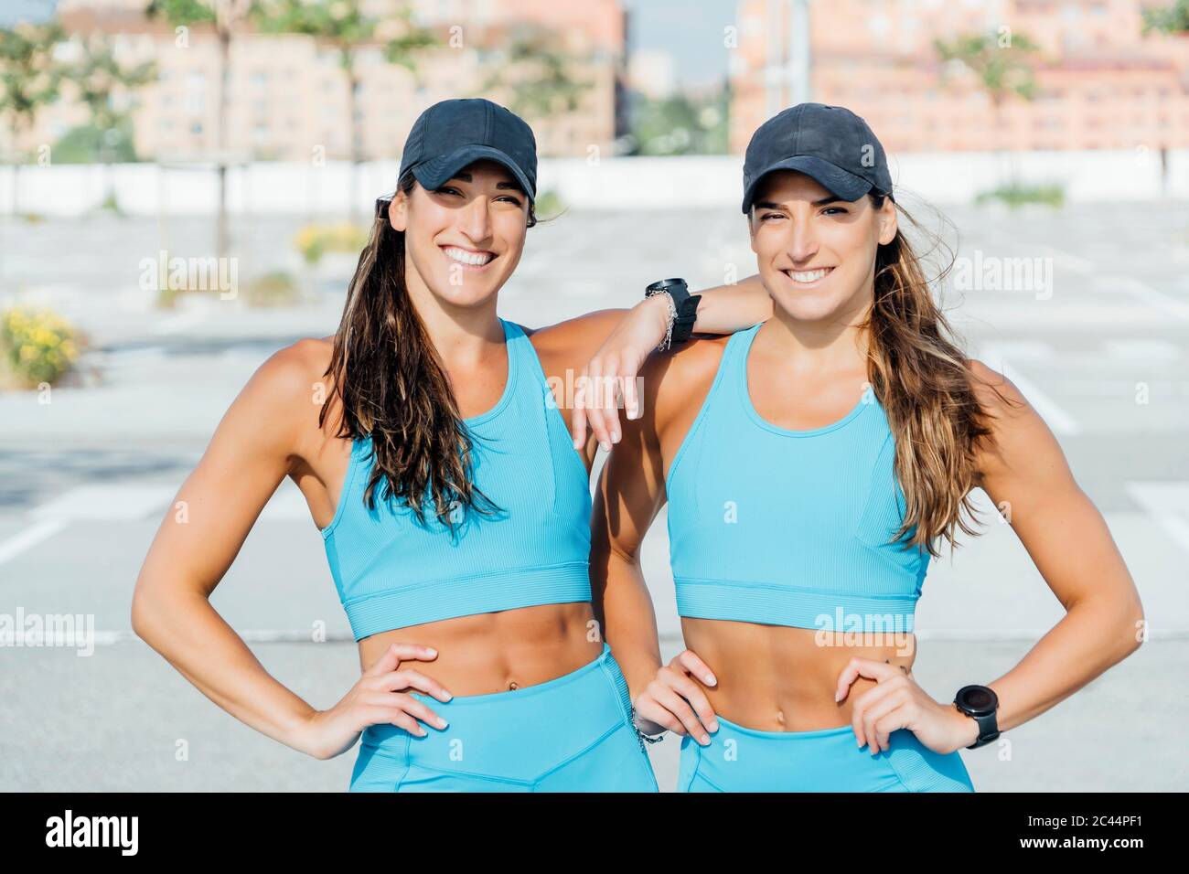 Retrato de hermanas gemelas jóvenes y confiadas en la sonrisa en ropa deportiva de pie al aire libre en el día soleado Foto de stock