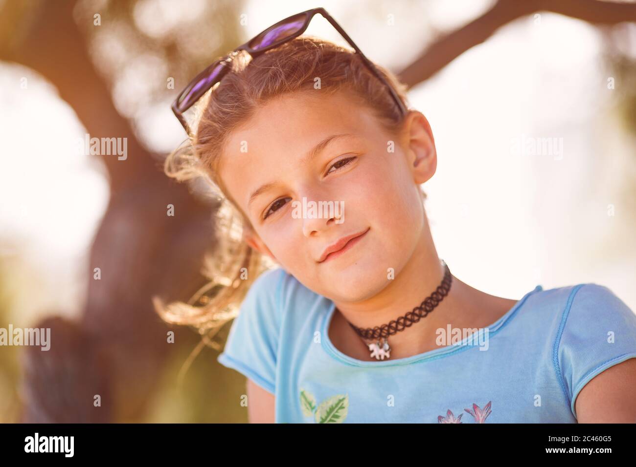 Niña, entre años, sentada en un árbol sonriendo a cámara con gafas de sol sobre la cabeza Foto de stock