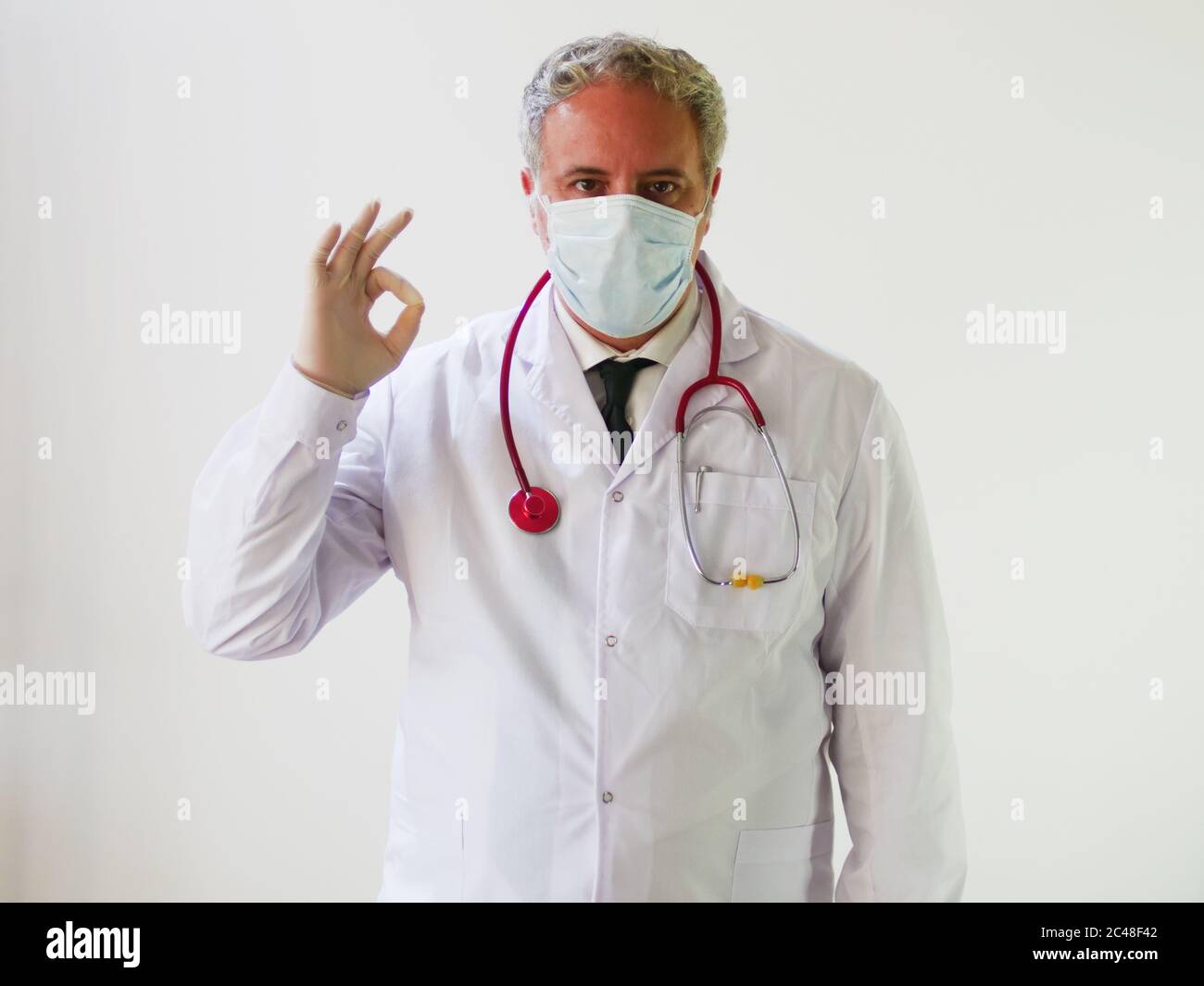 Hombre médico delantal con máscara médica protectora y guantes diciendo OK  con gesto de dedo en la clínica o Hospital Protección de la Salud debido  virus Corona COVID Fotografía de stock -