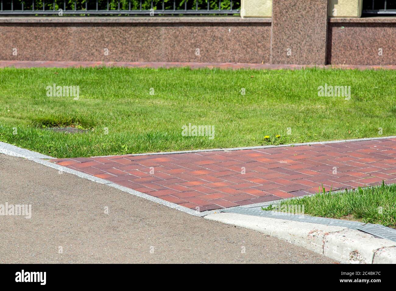 pavimento peatonal hecho de azulejos rojos que fluyen en la carretera de asfalto con un bordillo en la valla de piedra con un césped verde iluminado por la luz del sol. Foto de stock