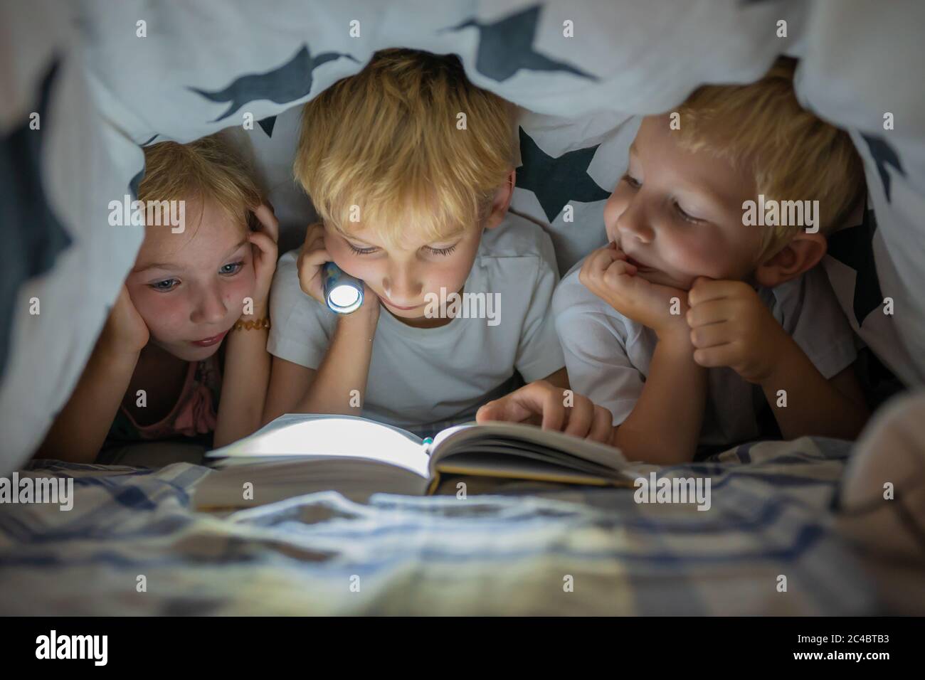 Tres niños pequeños están leyendo un libro con una linterna debajo de las cubiertas por la noche. Foto de stock