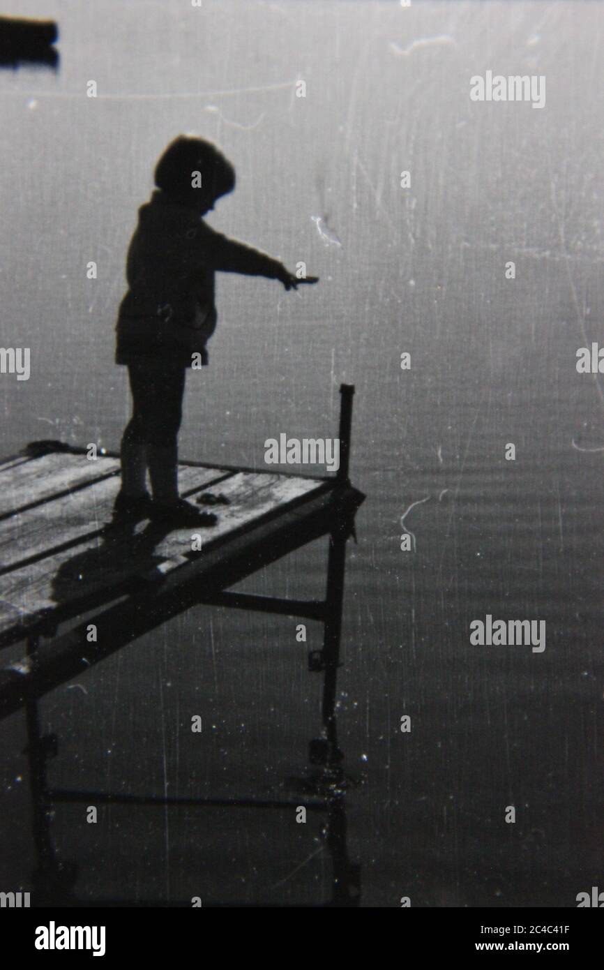 Foto en blanco y negro de un niño parado al final de un muelle mirando las aguas tranquilas. Foto de stock