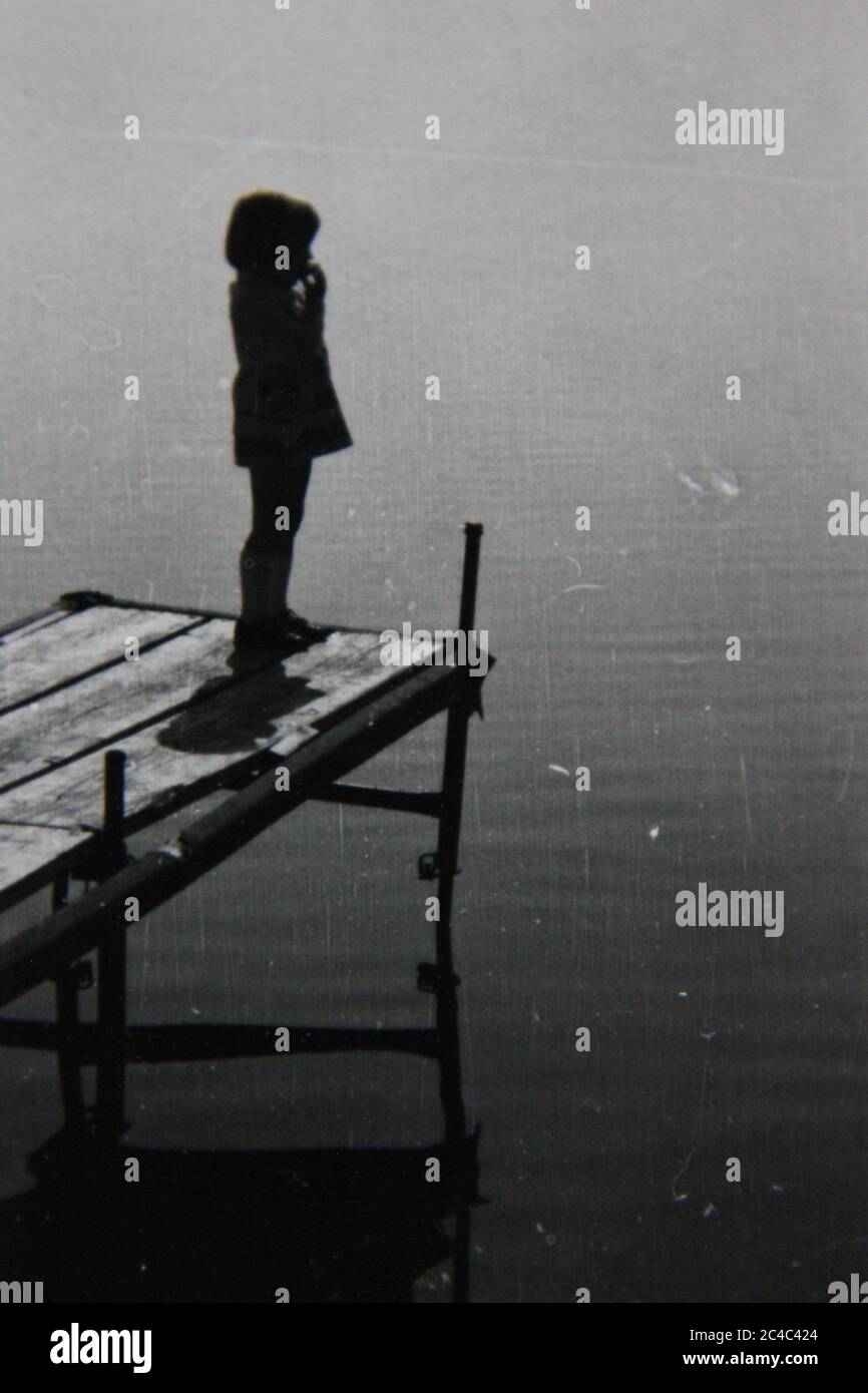 Foto en blanco y negro de un niño parado al final de un muelle mirando las aguas tranquilas. Foto de stock