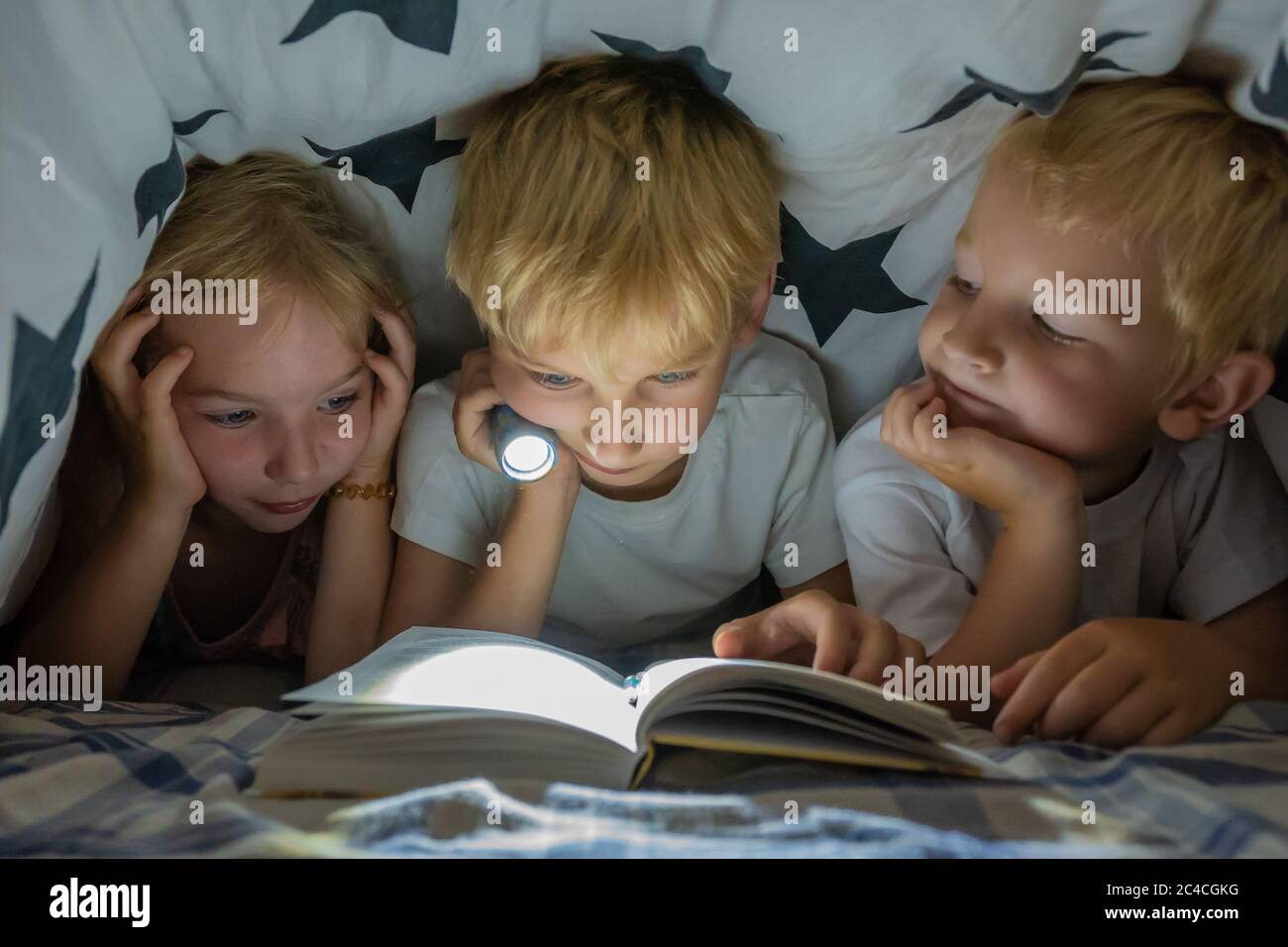 Tres niños pequeños están leyendo un libro con una linterna debajo de las cubiertas por la noche. Foto de stock