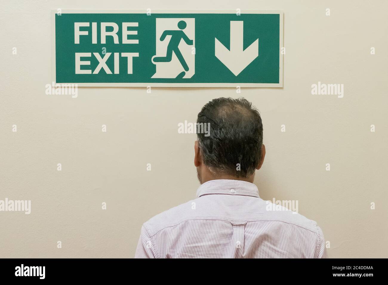 Hombre visto caminar en una pared sólida, debajo de un signo de salida de incendios situado en un complejo de oficinas. La salida de incendios está bajando las escaleras. Foto de stock