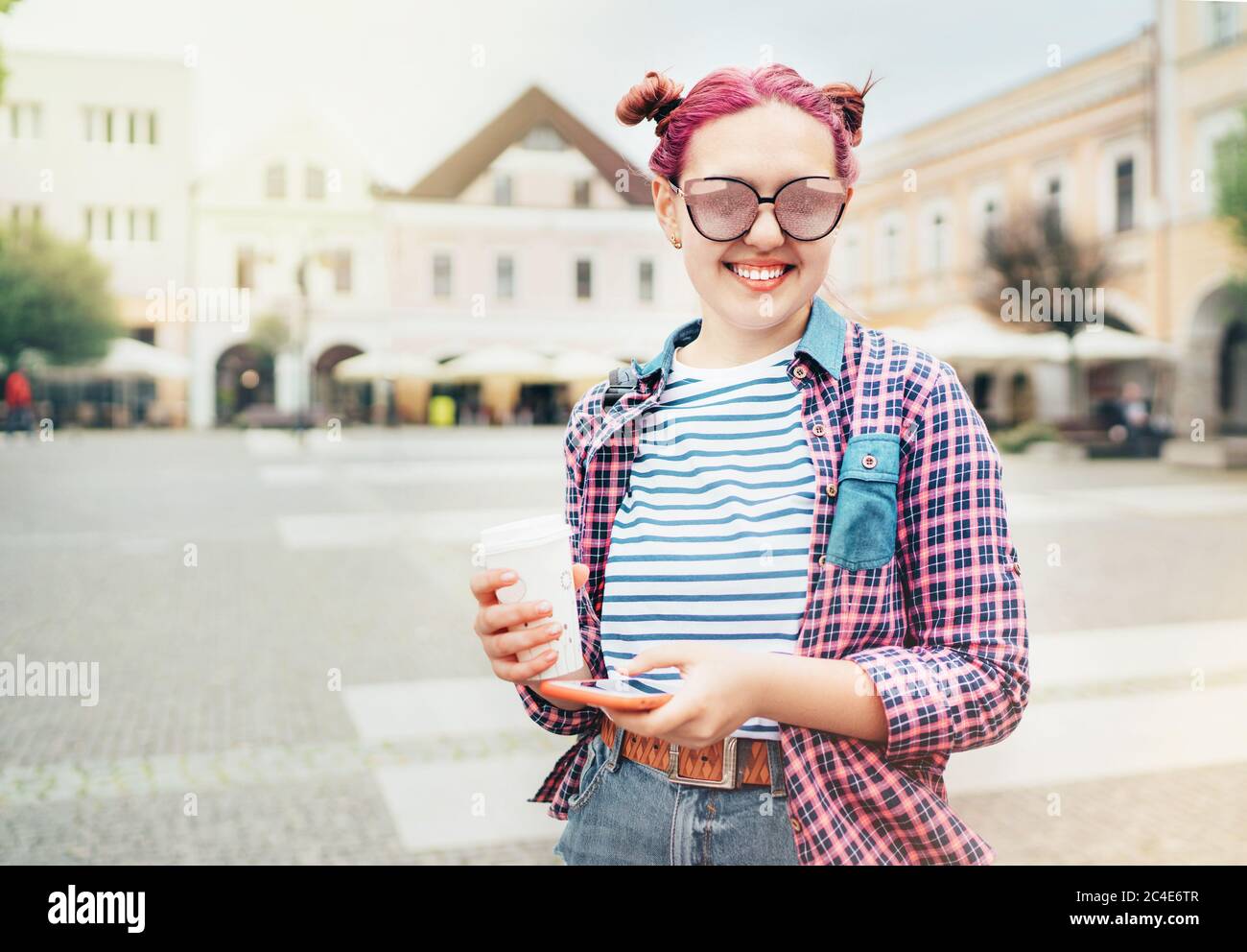 Retrato de la hermosa joven adolescente moderna con un extraordinario peinado en camisa de cuadros con casquillo 'café para llevar' y smartphone delgado. Moder Foto de stock