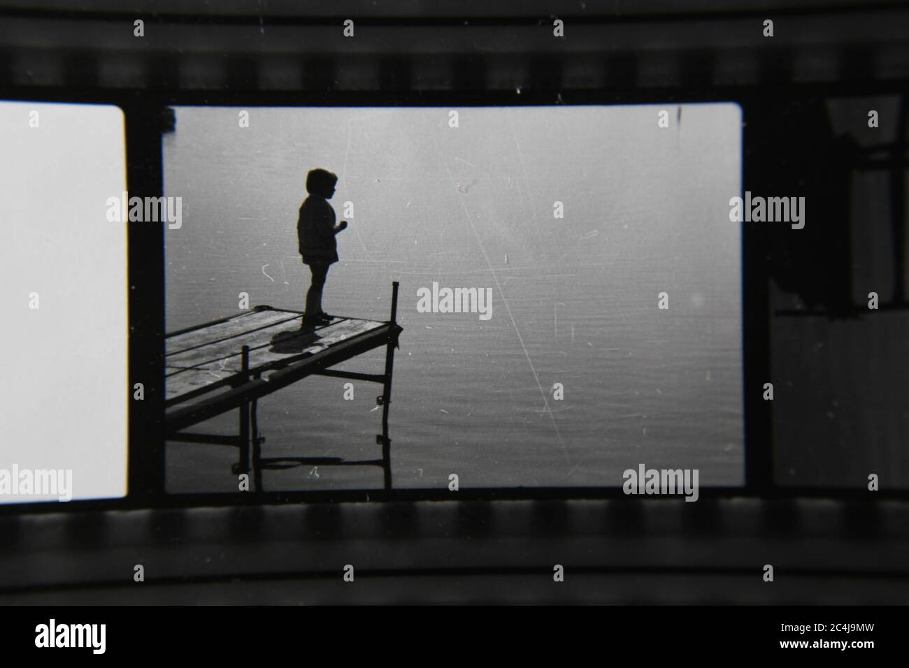 Foto en blanco y negro de un niño parado al final de un muelle mirando las aguas tranquilas. Foto de stock