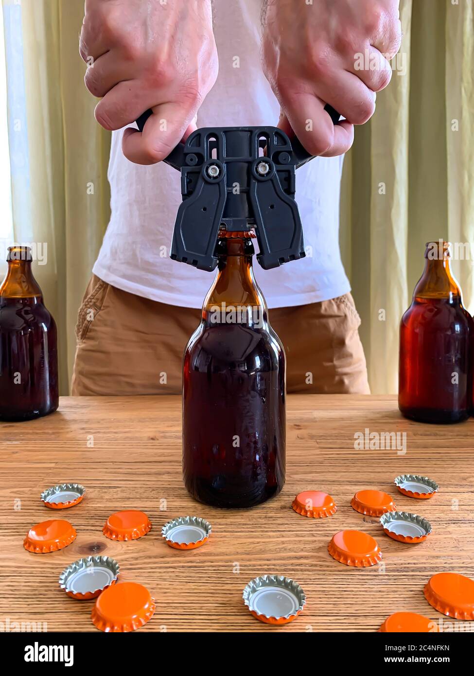 Cerveza artesanal en casa, el hombre cierra botellas de cerveza de vidrio  marrón con tapón de plástico en la mesa de madera con tapas de corona  naranja. Imagen vertical Fotografía de stock -