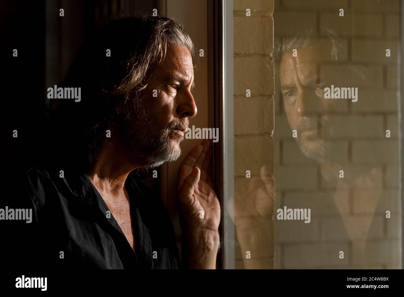 hombre en aislamiento propio, estancia concepto de casa, pensar el hombre reflexión en la ventana de cristal de la casa de ladrillo, preocupado de pelo largo hombre con barba, casa limitada Foto de stock