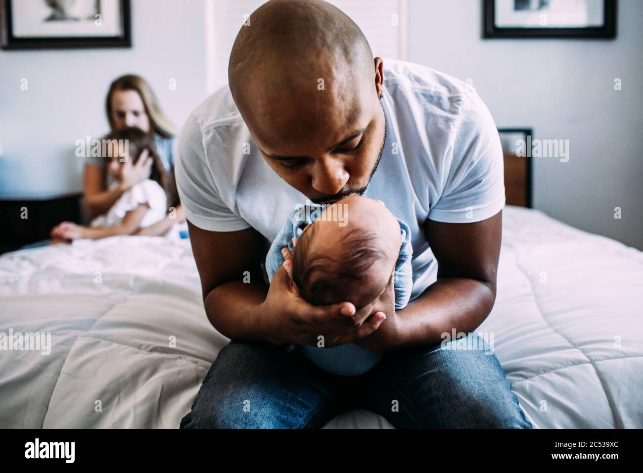 papá besando al recién nacido en la cama con la mamá y la hija adentro fondo Foto de stock