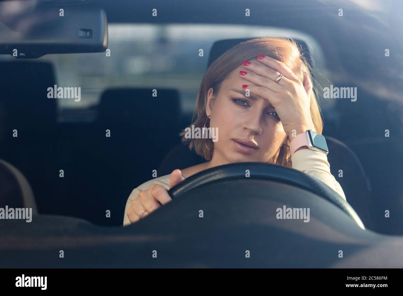 Exhausta joven mujer conductor sentado en su coche, sintiendo agotamiento emocional después del trabajo, mirando la cámara, tocando su frente. Salud mental, fatigu Foto de stock