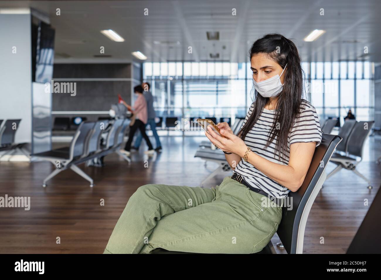 La máscara de protección de la mujer joven está sentada en un salón de espera y mirando su teléfono móvil en el aeropuerto. El virus de la corona se rompe. Foto de alta calidad Foto de stock