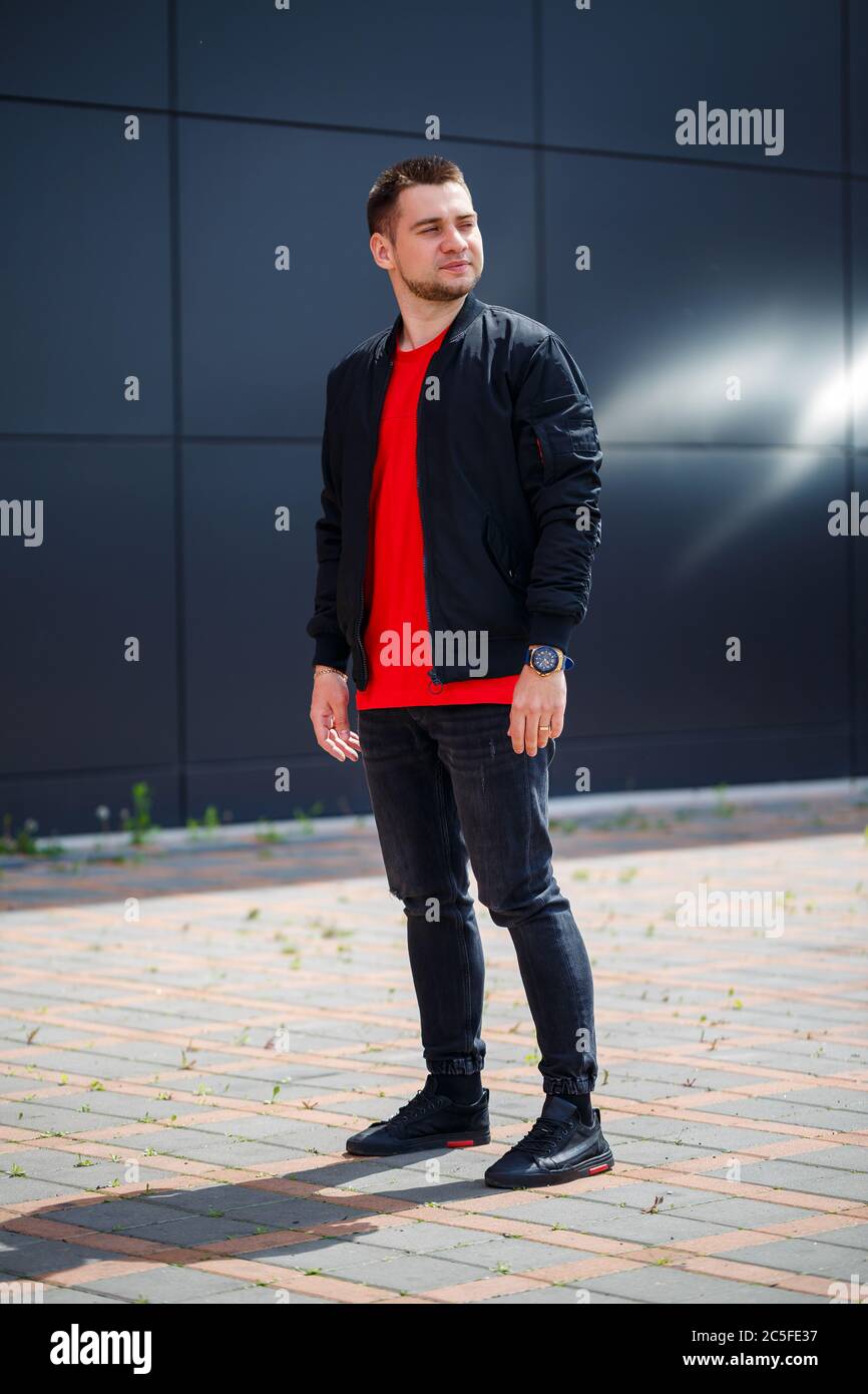 Un joven vestido de estilo informal, una chaqueta negra, jeans, una  camiseta roja. Está contra una pared gris Fotografía de stock - Alamy
