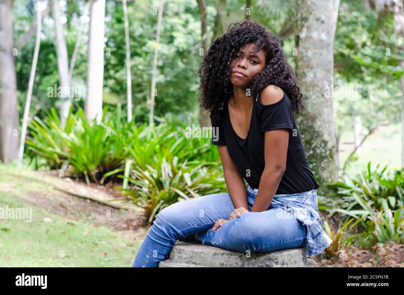 una joven negra de entre 20 y 30 años sentada leyendo un libro solo, en un parque, rodeado de árboles Foto de stock