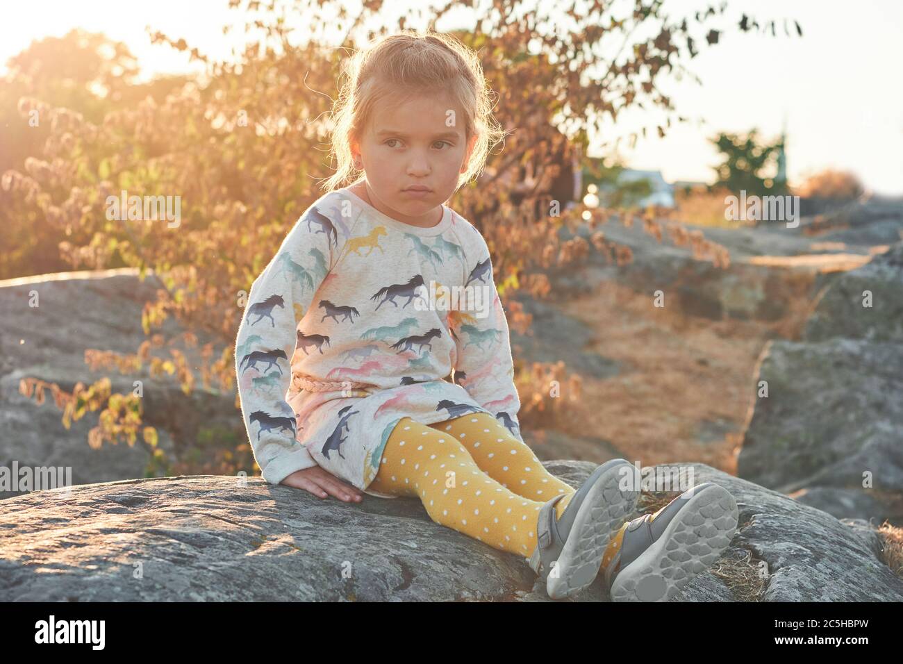 Chica triste sentada sobre piedras, puesta de sol sobre fondo Foto de stock