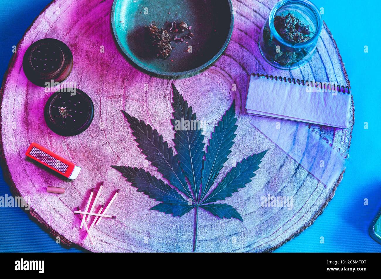 Molinillo con flores de cannabis trituradas, papeles ecológicos, cerillas, hoja seca de maleza sobre mesa de madera - enfoque en la hoja - luces de neón azules y púrpura edición Foto de stock