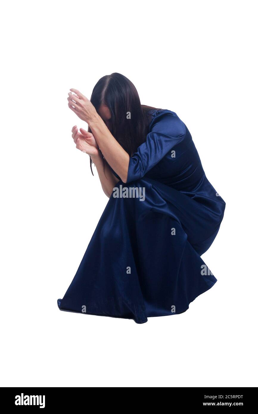 una morena sobre un fondo blanco aislado está en cuclillas, cubriendo su rostro con sus manos, en un clásico vestido azul Foto de stock