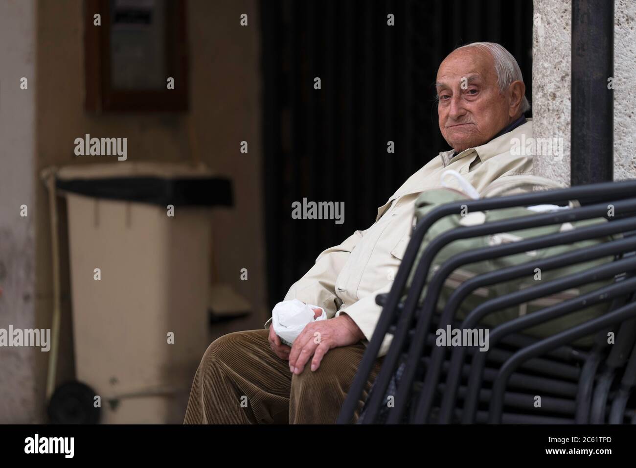 Viejo italiano con abrigo blanco se sienta en silla y mira en la cámara en la plaza Piazza del Popolo en Ascoli Piceno, Italia. Pila de otras sillas Foto de stock