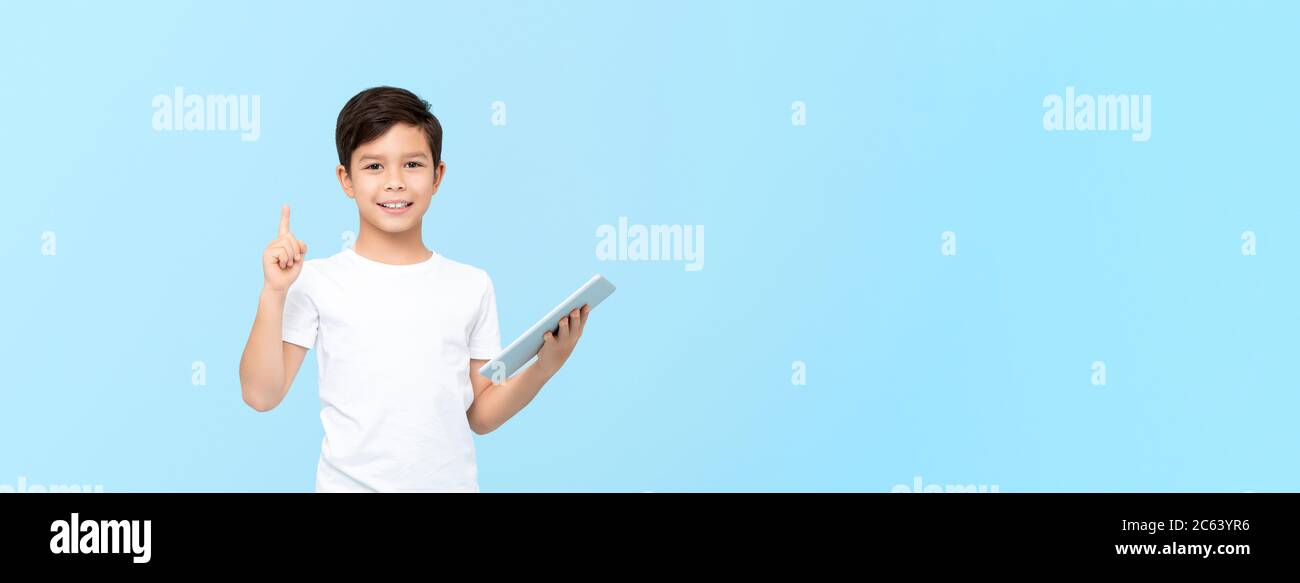Lindo chico de 10 años de edad mixto de carrera sosteniendo el ordenador de tableta con una mano apuntando hacia arriba al espacio vacío aislado en fondo azul claro banner Foto de stock