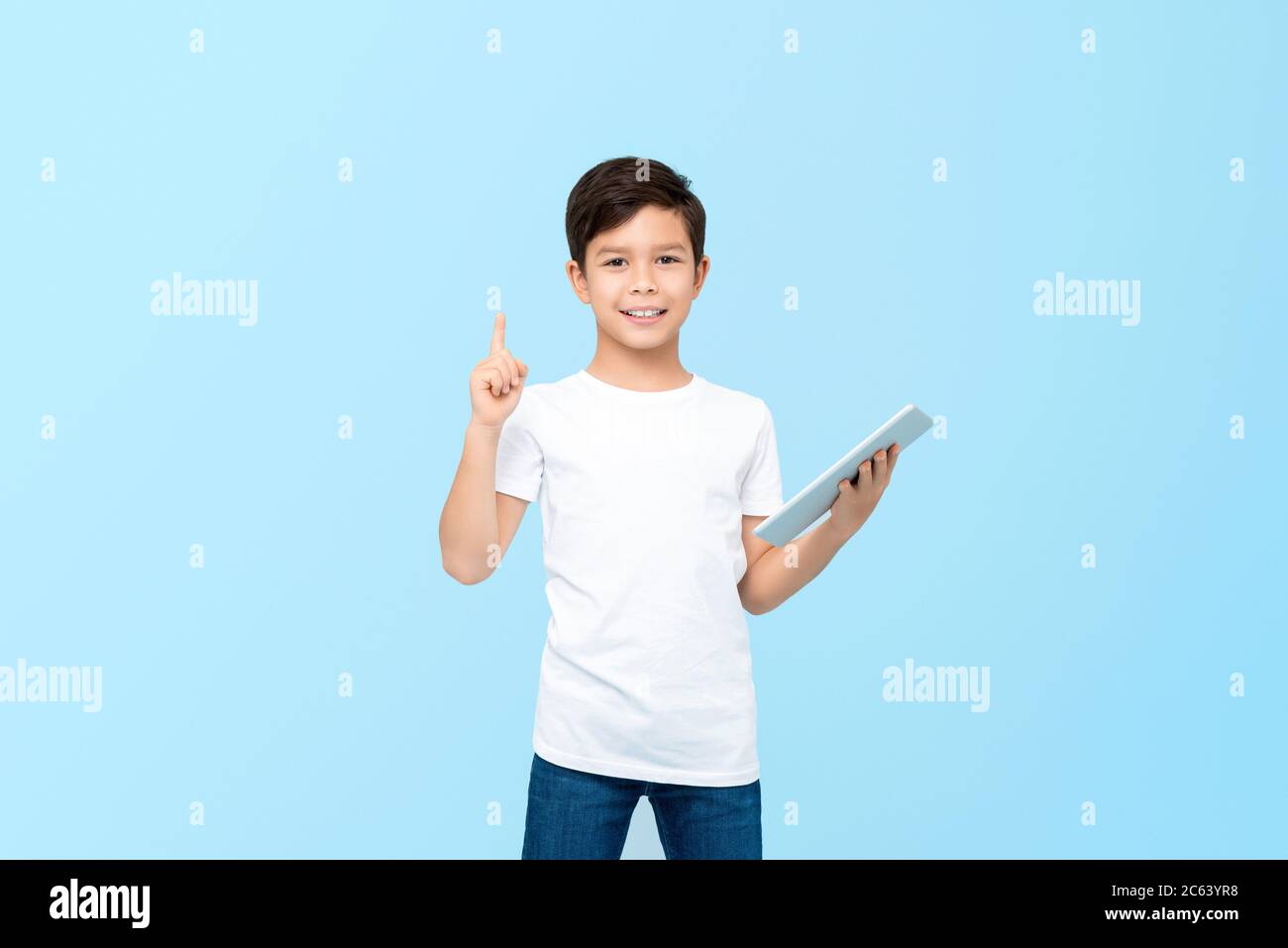 Lindo chico de 10 años de edad mixto de carrera sosteniendo el ordenador de tableta con una mano apuntando hacia arriba al espacio vacío aislado sobre fondo azul claro Foto de stock
