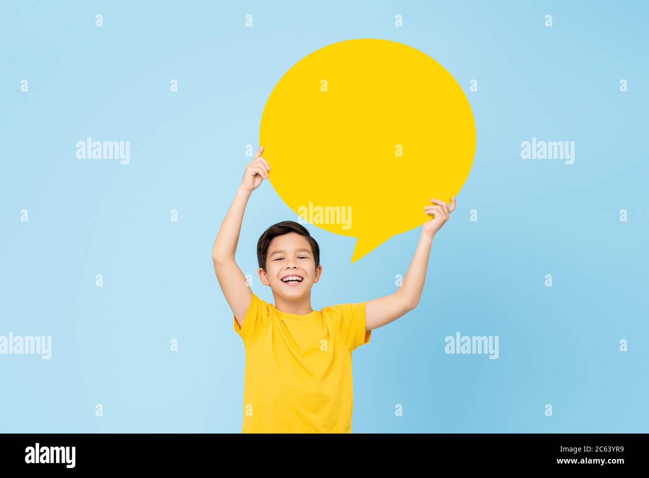 Retrato de cintura arriba de sonriente lindo niño asiático sosteniendo una burbuja de voz vacía en el fondo de estudio azul claro Foto de stock