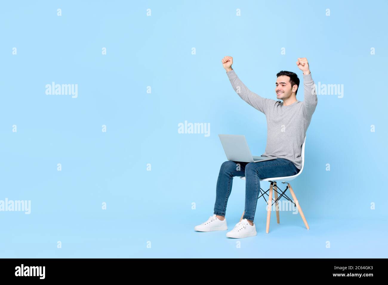 Un joven y ecstático que gana el hombre caucásico levantando ambos puños con ordenador portátil en el regazo contra fondo azul claro con espacio de copia Foto de stock
