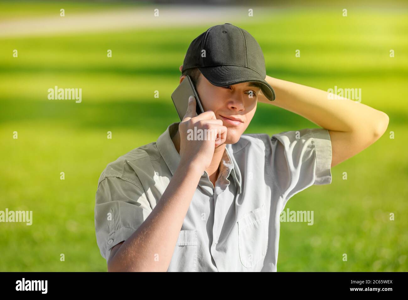 Joven adolescente de moda usando su teléfono inteligente o móvil mientras habla con un amigo mientras camina por un césped verde bajo la luz del sol con su mano a su p Foto de stock