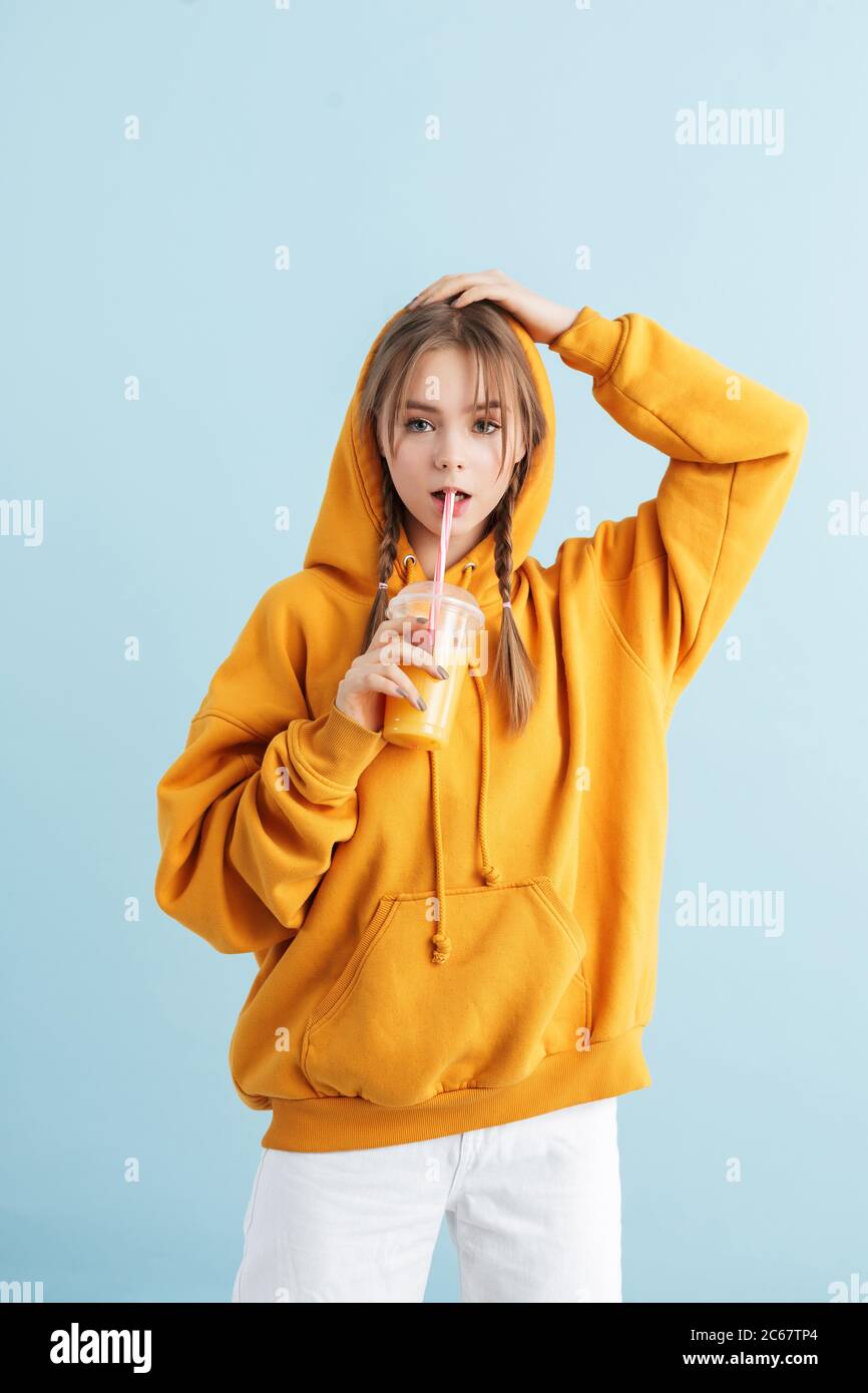 Joven hermosa chica con dos trenzas en naranja hoodie beber jugo en vaso de  plástico mientras que cuidadosamente mirando en la cámara sobre fondo azul  Fotografía de stock - Alamy