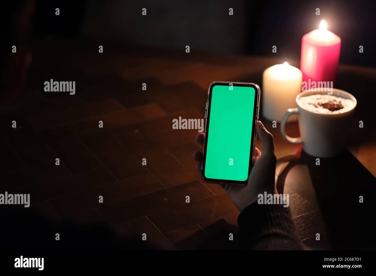vista en primera persona de la mano sosteniendo un smartphone con pantalla verde. Luces de velas borrosas y una taza de café en una mesa de madera. Fondo oscuro Foto de stock